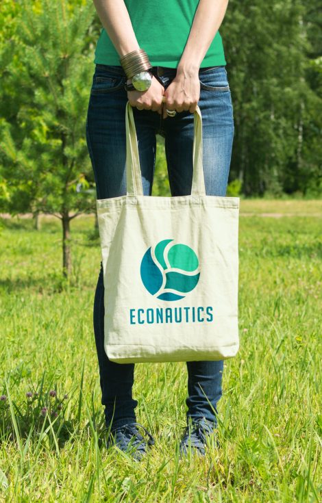 Woman standing in a grassy field in the sunshine, holding a reusable tote bag with the Econautics Sustainability Institute simplified flower logo
