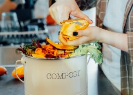 Compost the kitchen waste, recycling at home. Compost bin with vegetables cutted leftovers on the kitchen table with blurred woman cooking meal. Environmentally responsible, ecology. Selective focus.